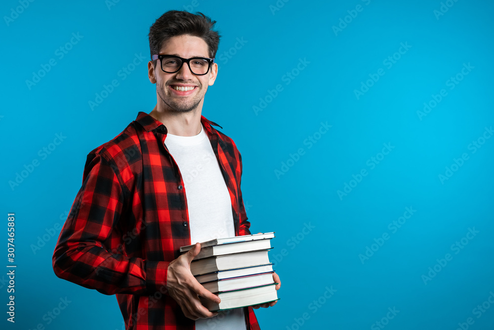 European student in red plaid shirt on blue background in studio holds stack of university books from library. Guy smiles, he is happy to graduate. - obrazy, fototapety, plakaty 