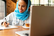 © Westend61 - Businesswoman wearing turquoise hijab in a cafe and writing in notebook