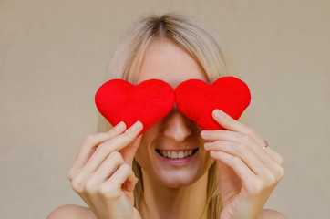 Lifestyle concept: attractive blonde woman with a radiant smile, having two small red hearts in her hands, closing her eyes with heart symbols, standing on a light background. Valentine's day