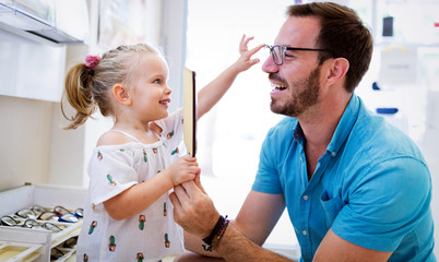 Wall Mural - Health care, eyesight and vision concept. Little girl choosing glasses with father at optics store