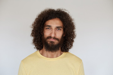 Indoor photo of pleasant looking positive young bearded man with brown curly hair smiling slightly while posing over white background, wearing casual clothes