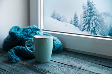 Winter background - cup with candy cane, woolen scarf and gloves on windowsill and winter scene outdoors. Still life with concept of spending winter time at cozy home with cold weather outdoors