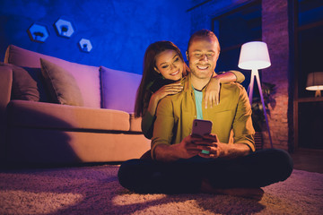 Wall Mural - Portrait of his he her she nice attractive lovely glad cheerful cheery married spouses sitting on carpet spending free time using digital device app 5g at night home house apartment