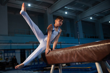 Wall Mural - Little male gymnast training in gym, flexible and active. Caucasian fit little boy, athlete in white sportswear practicing in exercises for strength, balance. Movement, action, motion, dynamic concept