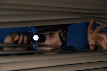 Wall Mural - Male security guard with flashlight looking through window blinds in darkness