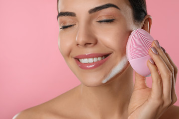 Poster - Young woman using facial cleansing brush on pink background, closeup. Washing accessory