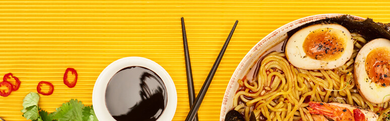 Wall Mural - top view of seafood ramen near fresh ingredients, soy sauce and chopsticks on yellow surface, panoramic shot