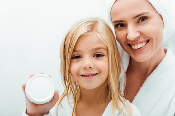 cute kid holding container with cosmetic cream near cheerful mother in bathrobe