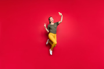 Poster - Full body photo of crazy funny lady jumping high making selfies showing v-sign symbol cheerful mood wear casual yellow pants green t-shirt isolated red color background