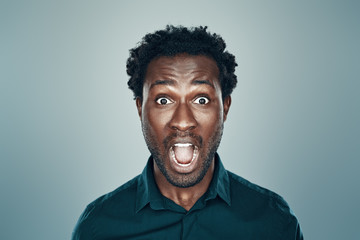 Surprised young African man looking at camera and keeping mouth open while standing against grey background