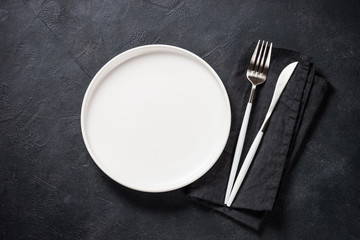 White plate, cutlery and napkin on black table top view.