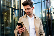 Young Stylish Man In Trench Coat Using Cellphone On Street