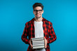 © kohanova1991 - European student in red plaid shirt on blue background in studio holds stack of university books from library. Guy smiles, he is happy to graduate.