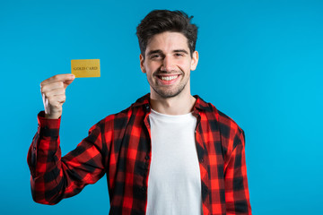 Successful man in plaid shirt showing unlimited gold credit card on blue studio background. Student, money concept