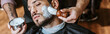 Panoramic shot of barber applying shaving cream on face of handsome bearded man
