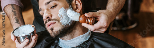 Fotografía panoramic shot of barber applying shaving cream on face of handsome bearded man
