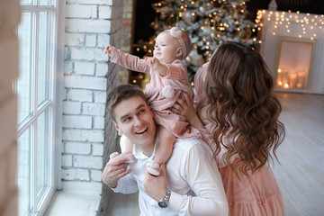 A happy family, father, mother and little daughter, spend time together on Christmas evening in a bright room indoors against a background of a beautifully decorated Christmas tree with garlands and g