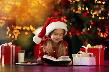 Poster - Little girl in Santa Claus cap reading book near Christmas tree indoors