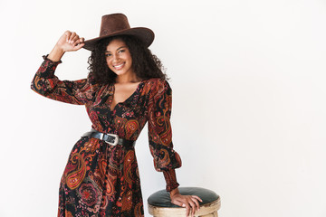 Wall Mural - Image of young african american cowgirl in dress and hat standing by chair