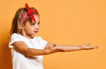 Wall Mural - Nice kid girl in white t-shirt and red headband is holding something on her open palms on yellow with copy space