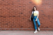 © Vasya - Attractive serious young brunette girl in blue jeans and yellow t-shirt, with black shoulder bag isolated over red brick wall