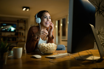 Young woman having fun while eating popcorn and watching movie on a computer in the evening.