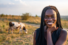 Close Up Portrait Of The Young African Girl In Black Vest Among The Field, Cow Graze On The Background