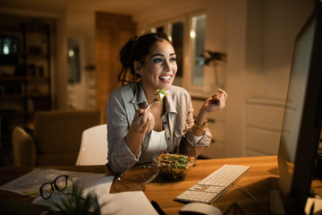 Young woman eating salad while working on a computer late at night.