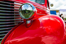 Old Vintage American Bright Red Pickup Car Front Side Close Up View, With Chrome Glass Headlights Light Lamp Parts And Grille During An Outdoor Show
