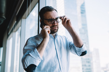 Wall Mural - Caucasian employer in classic spectacles for provide eyes protection using cellphone gadget for making service consultation during work day, puzzled businessman calling to partner via cellular app