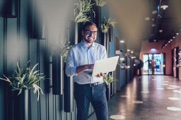 Wall Mural - Half length portrait of successful boss using netbook technology with 4g wireless internet connection in workspace interior, prosperous employee in classic eyewear holding laptop computer and smiling