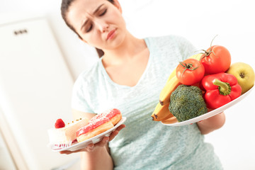 Canvas Print - Body Care. Chubby girl standing in kitchen choosing between healthy and unhealthy food close-up blurred background