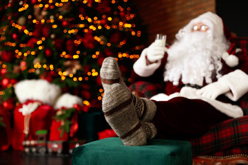 Poster - Santa Claus with glass of milk resting in armchair near Christmas tree, focus on legs