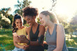 © StratfordProductions - Smiling female diverse friends looking at mobile phone smiling and having fun in the morning at the park - friends laughing together