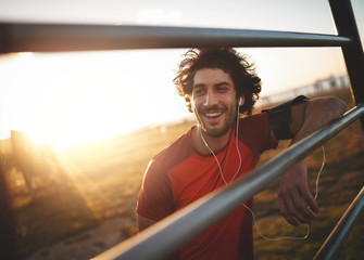 Portrait of a smiling young male athlete with earphones in his ears leaning on horizontal bars resting after run