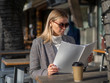 Fashion woman in sunglasses and a coat on a cold spring day sitting in a street cafe and reading a magazine with empty cover with copy space for your mockup