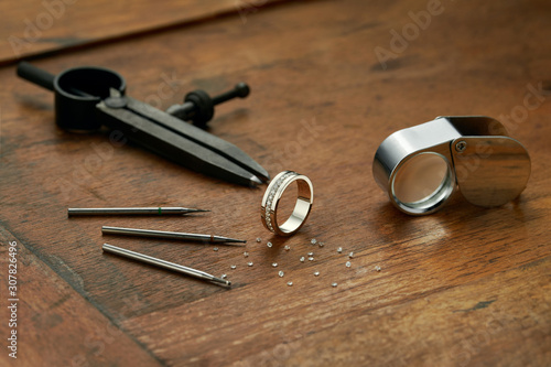 Workplace of a jeweler with tools and equipment for working on an old wooden table. Ready-made diamond engagement ring, compass, diamonds, magnifying glass. Engraver at work on jewelry from diamonds