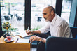 © BullRun - Old male using tablet siting while writing in notepad at table in cafe