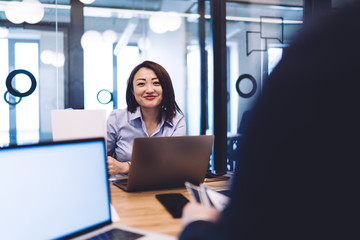 Smiling ethnic woman working at laptop and looking at camera in office