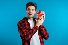 Handsome Man Holding Gift Box On Blue Studio Background Smiles To Camera. Happy European Guy Received Present And Interested In What's Inside.