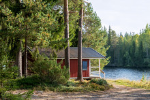 Red Wooden Finnish Traditional Cabins Cottages In Green Pine Forest Near River. Rural Architecture Of Northern Europe. Wooden Houses In Camping On Sunny Summer Day
