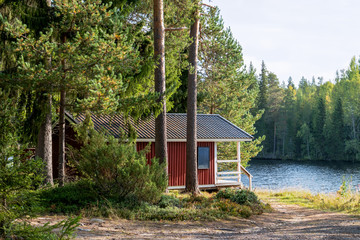 Red wooden finnish traditional cabins cottages in green pine forest near river. Rural architecture of northern Europe. Wooden houses in camping on sunny summer day