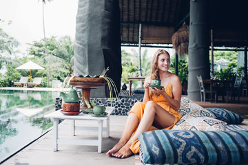 Cheerful woman drinking tea next to pool in hotel