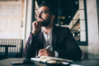 Dreamy caucasian bearded business owner looking away thinking about future working in his office, pensive formally dressed male entrepreneur pondering on successful strategy making notes in notepad