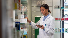An Young Female Pharmacist Consultant Is Checking With A Tablet Medicines On The Shelf Of Drug Store. 