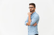 © Mangostar - Serious pensive guy touching chin and looking at camera. Handsome young man in casual shirt and glasses standing isolated over white background. Advertising concept