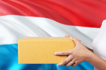 Luxembourg delivery service. International shipment theme. Woman courier hand holding brown box isolated on national flag background.