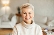 canvas print picture - Close up image of happy good looking elegant fifty year old woman wearing warm cozy jumper, pearl earrings and short stylish hairdo being in good mood sitting in living room, smiling broadly at camera