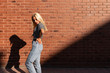 © Vasya - Side view portrait of young blonde girl in gey t-shirt and jeans that syands in the sun, holds her hands on her waist, isolated over red brick wall