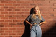 © Vasya - Serious blonde girl in grey t-shirt and jeans stands in the sun with hands on her waist isolated over red brick wall